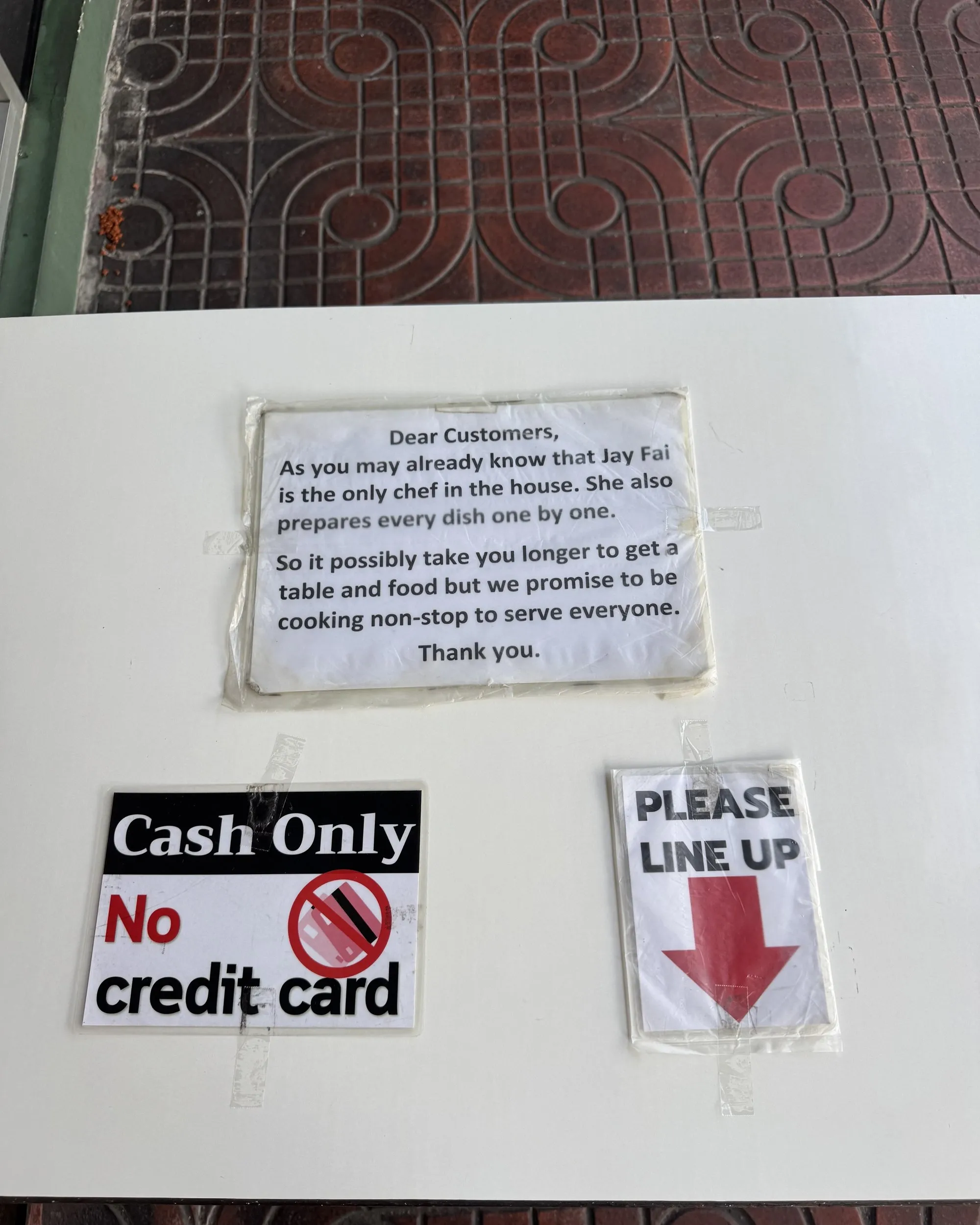 Un tavolo bianco sul marciapiede con tre cartelli fissati con il nastro adesivo: un avviso ai clienti che spiega che Jay Fai è l'unica cuoca e prepara ogni piatto uno alla volta, un cartello Cash Only, No credit card e un cartello Please line up con una freccia rossa.