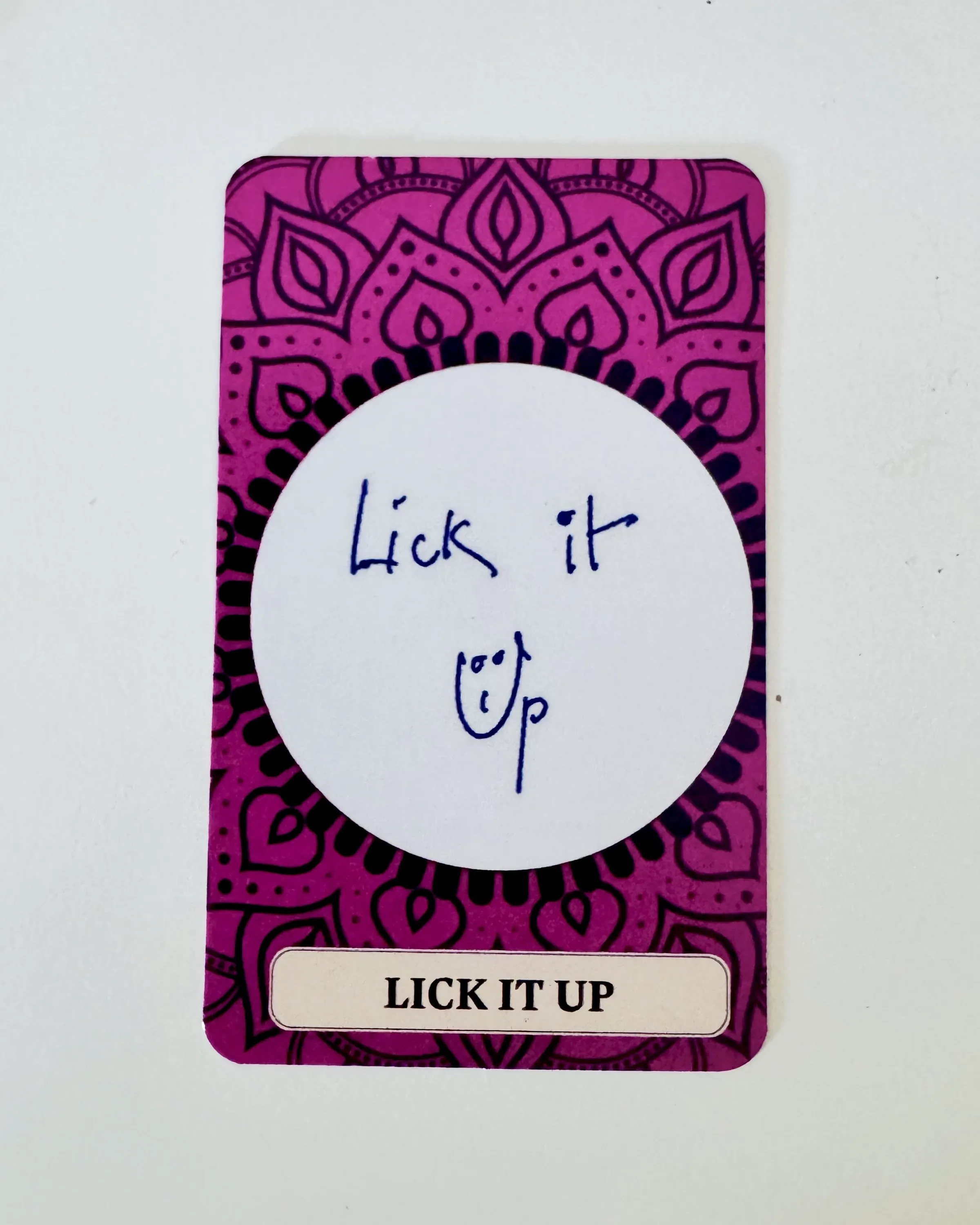 Una carta con cornice decorativa viola: al centro, scritto a mano a penna, Lick it Up con un piccolo volto sorridente; in basso l'etichetta stampata LICK IT UP.