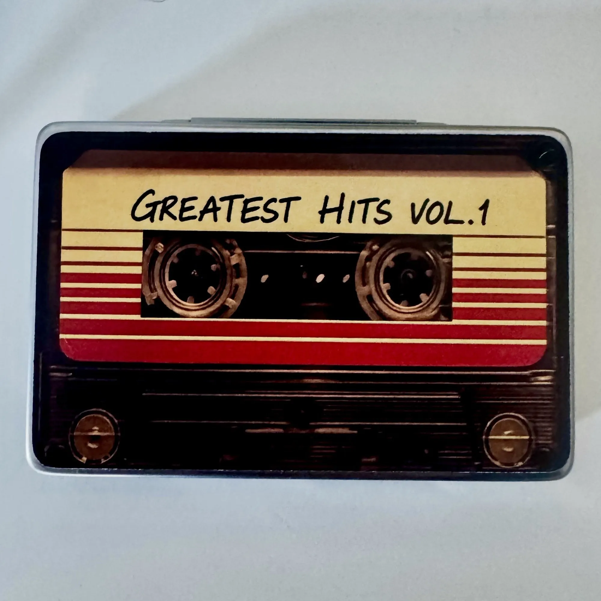 Una scatola rettangolare di alluminio decorata come una musicassetta, con l'etichetta Greatest Hits Vol. 1.